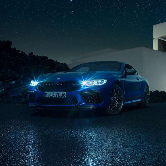 BMW M8 Coupé, three-quarter front view. BMW M8 Coupé in Marina Bay Blue metallic with shining LED headlights, standing three-quarter front view in front of villa at night.