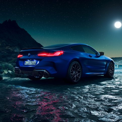 BMW M8 Coupé, three-quarter rear view. BMW M8 Coupé in Marina Bay Blue metallic with illuminated LED rear lights, standing three-quarter rear view with full moon.