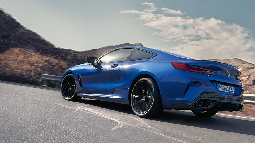 BMW 8 Series Coupé G15 LCI Facelift three-quarter rear view driving BMW 8 Series Coupé M Portimao Blue metallic G15 LCI Facelift three-quarter rear view driving in front of hills