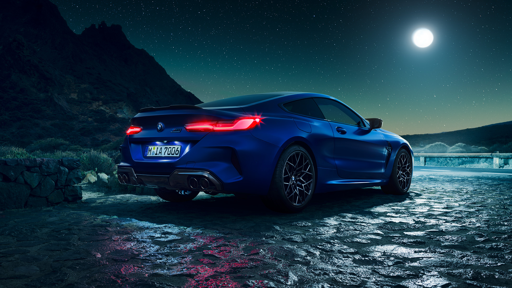 BMW M8 Coupé, three-quarter rear view. BMW M8 Coupé in Marina Bay Blue metallic with illuminated LED rear lights, standing three-quarter rear view with full moon.