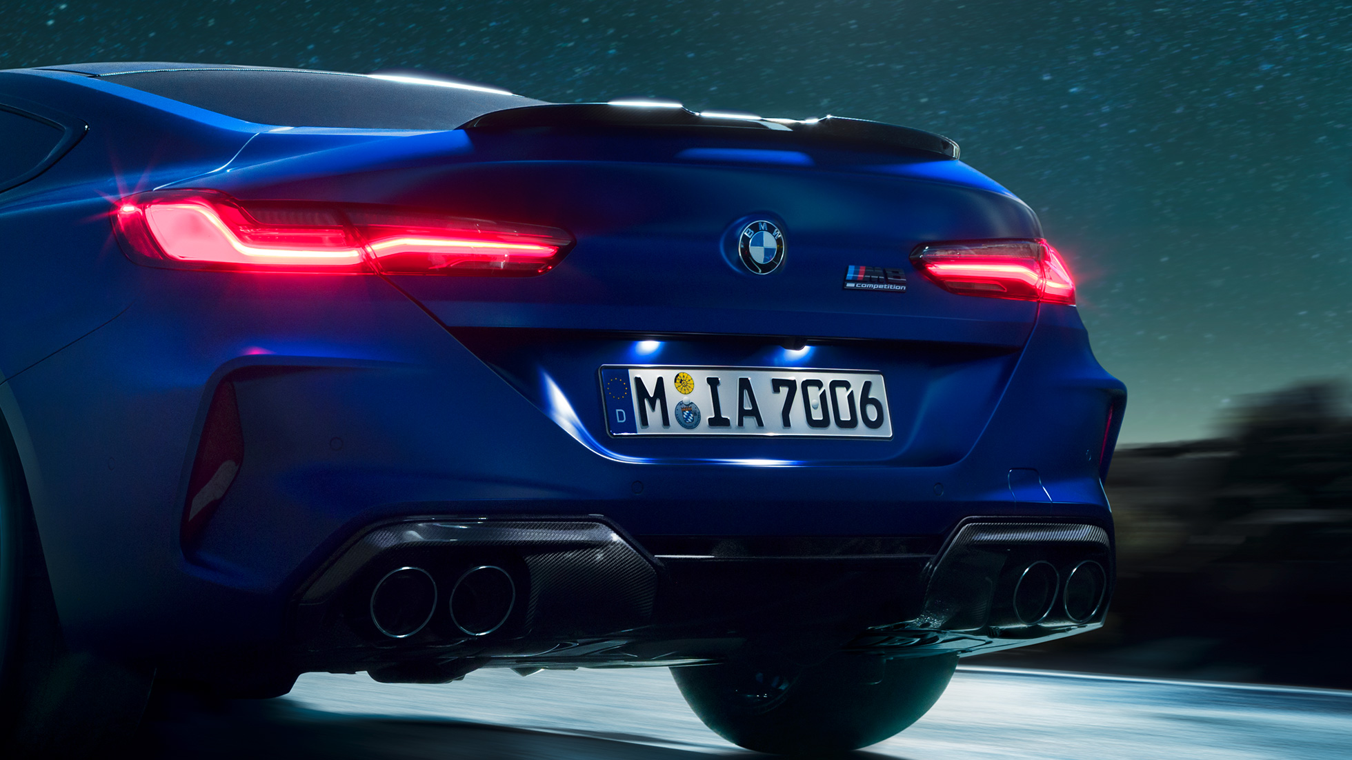 M diffuser in Carbon Black. BMW M8 Coupé in Marina Bay Blue metallic, close-up rear at night with M diffuser in Carbon Black.