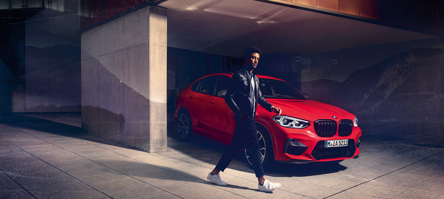 BMW X4 M Competition, three-quarter side view. BMW X4 M Competition in Toronto Red metallic, exterior, three-quarter side view with young man.