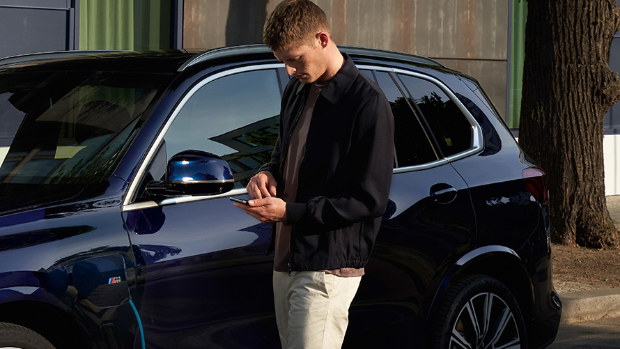 Man in front of BMW looking at his smartphone Remote Software Upgrade Man in front of BMW looks at his smartphone